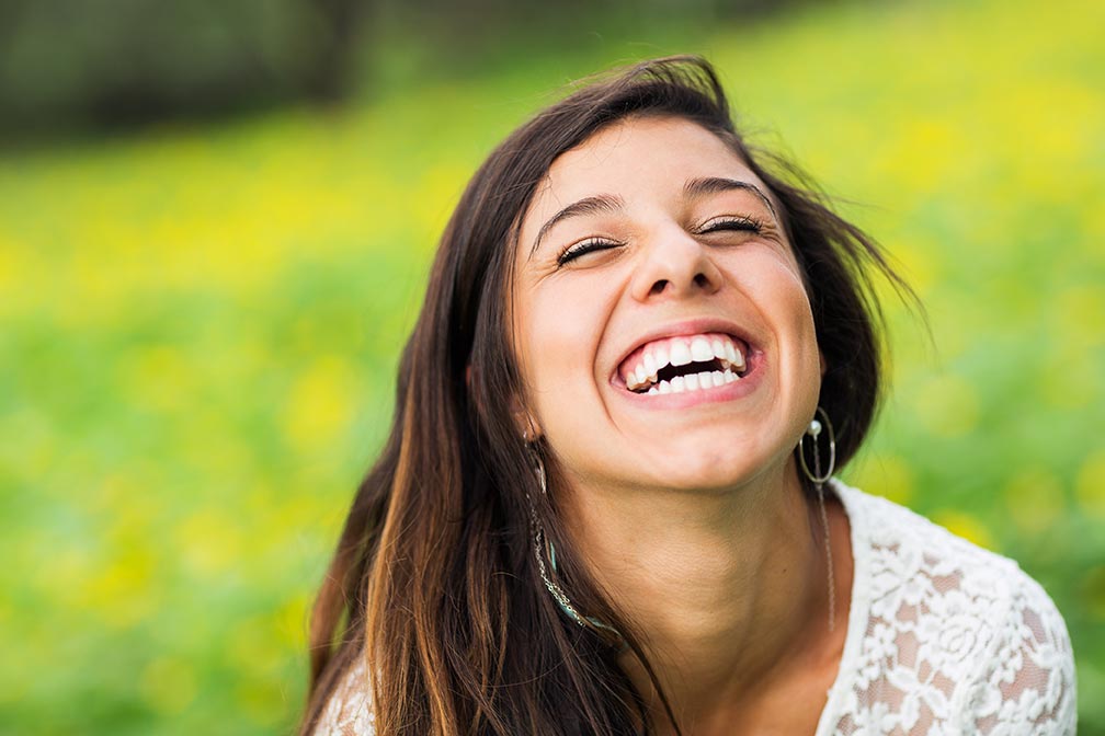 A young woman with white, healthy teeth sits outdoors on the grass and laughs happily.